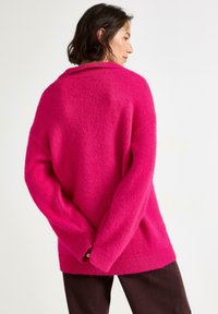 Uldet rosa oversized sweater med sjalskrave og ribbede manchetter og kant; båret med mørkebrune bukser, der viser en afslappet pasform.
