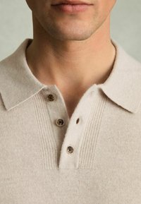 Polo en tricot beige avec une patte de boutonnage côtelée texturée, trois boutons marron et un col structuré, présenté de la poitrine vers le haut.