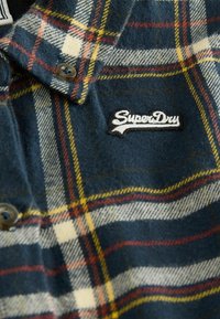 Mörk marinblå flanellskjorta med rutigt mönster i röd, gul och vit. Inkluderar en logotypapplikation i svart och vitt samt knapp.