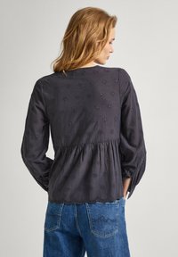 Blusa ricamata grigio scuro con taglio peplum, maniche lunghe e orlo scalloppato. Presenta motivi floreali decorativi e tessuto testurizzato.