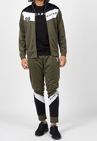Olivově zelený teplákový komplet s černými a bílými akcenty, zipová bunda a zúžené kalhoty se diagonálním blokováním barev a detaily loga.