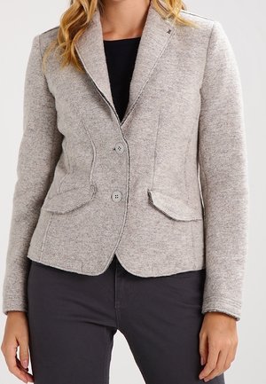 Femme portant un blazer gris clair à deux boutons avec des poches à rabat, sur un haut noir et un pantalon foncé.