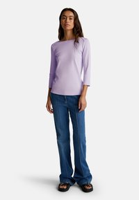 Donna in piedi con una maglietta a maniche lunghe color lavanda, jeans blu con cuciture verticali e sandali neri con punta aperta, su uno sfondo bianco.