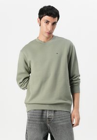 Olijfgroene sweatshirt met lange mouwen, ronde hals en ribgebreide manchetten. Heeft een klein logo op de borst. Gepaard met vervaagde grijze jeans.