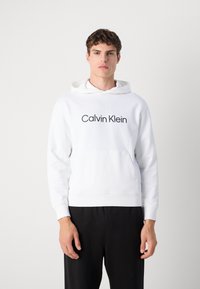 Hanorac alb din bumbac, cu buzunar frontal, mansete și tiv cu dungi, având logo-ul "Calvin Klein" în text îngroșat negru pe piept.