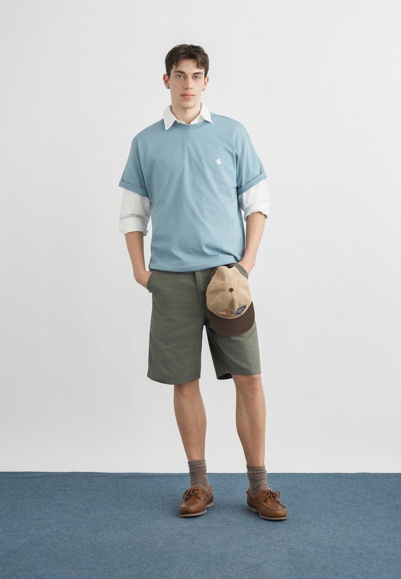 Jeune homme debout, les mains dans les poches tenant une casquette beige et marron, portant un T-shirt bleu clair par-dessus une chemise blanche à manches longues, un short vert et des chaussures marron.