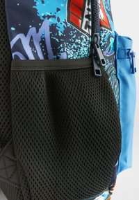 La mochila cuenta con un bolsillo de malla negra, un acento de tela azul y un diseño gráfico multicolor con salpicaduras de azul y púrpura.
