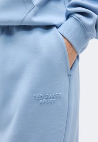 Σετ ανοιχτό μπλε φούτερ και παντελονιού με χέρι στην τσέπη, δείχνοντας το ανάγλυφο λογότυπο "Ted Baker Sport" στο παντελόνι κοντά στην τσέπη.