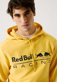 Felpa gialla in morbido tessuto, dotata di tasca a marsupio, coulisse regolabile e logo stampato "Red Bull Racing" con dettagli neri.