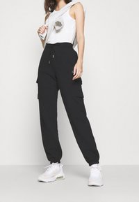 Černé cargo kalhoty s elastickým pasem a šňůrkou. Kombinace s bílým tílkem a šedým svetrem přehozeným přes rameno.