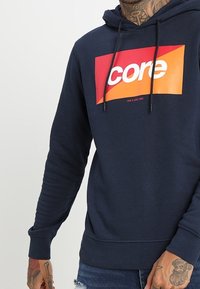 Sudadera azul marino hecha de tela suave, con un logotipo rectangular "core" colorido en rojo, naranja y blanco. Detalle de capucha con cordón.