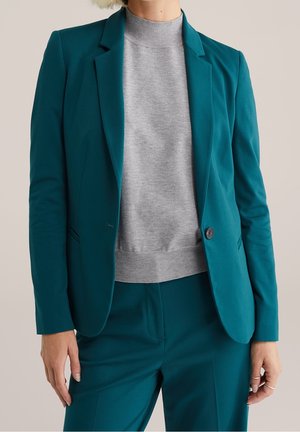 Persoon die een op maat gemaakte teal blazer en bijpassende broek draagt met een lichtgrijze trui met ronde hals, staand tegen een neutrale achtergrond.