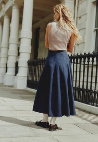 Tamsiai mėlyna džinsinė maksi sijonas su aukštu juosmeniu ir klostėmis, derinamas su smėlio spalvos marškinėliais be rankovių. Matomi rudi batai ir banguotos baltos kojinės.