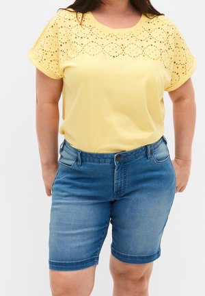 Top de algodón amarillo con detalles de ojal en los hombros, combinado con pantalones cortos de mezclilla azules. Los pantalones cortos cuentan con un diseño de botones en la parte delantera y bolsillos.