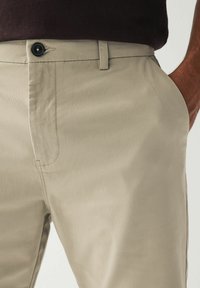 Pantalones beige de tejido suave, con cierre de botón, bolsillos y un ajuste entallado con detalles de costura sutiles.