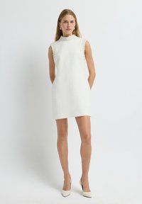 Witte mouwloze jurk met hoge kraag, gestructureerde stof en parelaccenten op de schouders. Gestyled met spitse beige hakken.