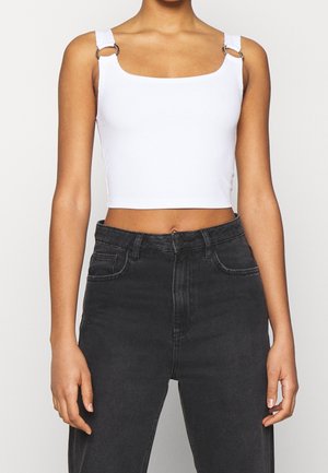 Persona que lleva una camiseta blanca corta con detalles en anillo en las tiras y jeans negros de cintura alta, contra un fondo liso.