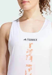 Witte sportieve tanktop met een ronde hals, oranje gestreepte grafische details en het "TERREX" logo in het zwart op de bovenste borstpartij.