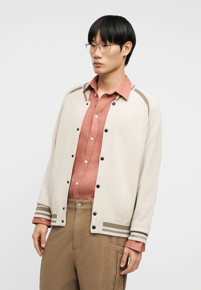 Νέος άνδρας με κοντά μαύρα μαλλιά και γυαλιά, φοράει ένα ανοιχτό μπεζ σακάκι με κουμπιά πάνω από ένα σκουροκόκκινο πουκάμισο και μπεζ παντελόνι, κοιτάζει προς τα πλάγια.