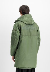 Giacca imbottita verde oliva con cappuccio, caratterizzata da un grande logo sul retro, tasca con zip sul maniche sinistro e una texture liscia e elegante.