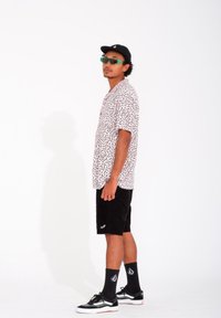 Kurzärmliges, weißes Hemd mit schwarzem abstraktem Muster, schwarze Shorts, schwarze Turnschuhe, grüne Sonnenbrille und schwarze Mütze, rückwärts getragen.