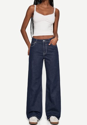 Jeans denim blu scuro a gamba larga con cuciture visibili, tasche frontali e chiusura con bottone, abbinati a un top bianco aderente.