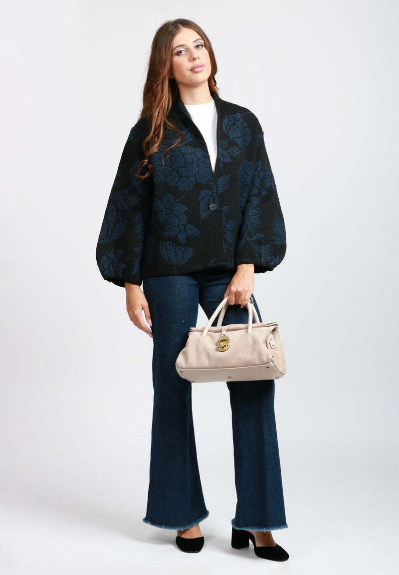 Cardigan oversize nero con motivo floreale e chiusura con bottoni, abbinato a jeans svasati blu e una borsa beige chiaro. Scarpe nere completano l'outfit.