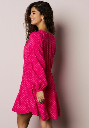 Roosa täpiline kleit, millel on voolav disain, paftvarrukad ja kogutud vöökoht, hemline avaneb laiuvaks. Tekstureeritud kangas.