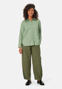 Lichtgroene lange mouw knoopshirt met een borstzak, gecombineerd met losse olijfgroene broek en zwarte schoenen. Gladde stoftextuur.