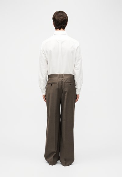 Άνδρας που στέκεται με την πλάτη, φοράει λευκό πουκάμισο με μακριά μανίκια και καφέ παντελόνι με φαρδιές πιέτες και πίσω τσέπες.