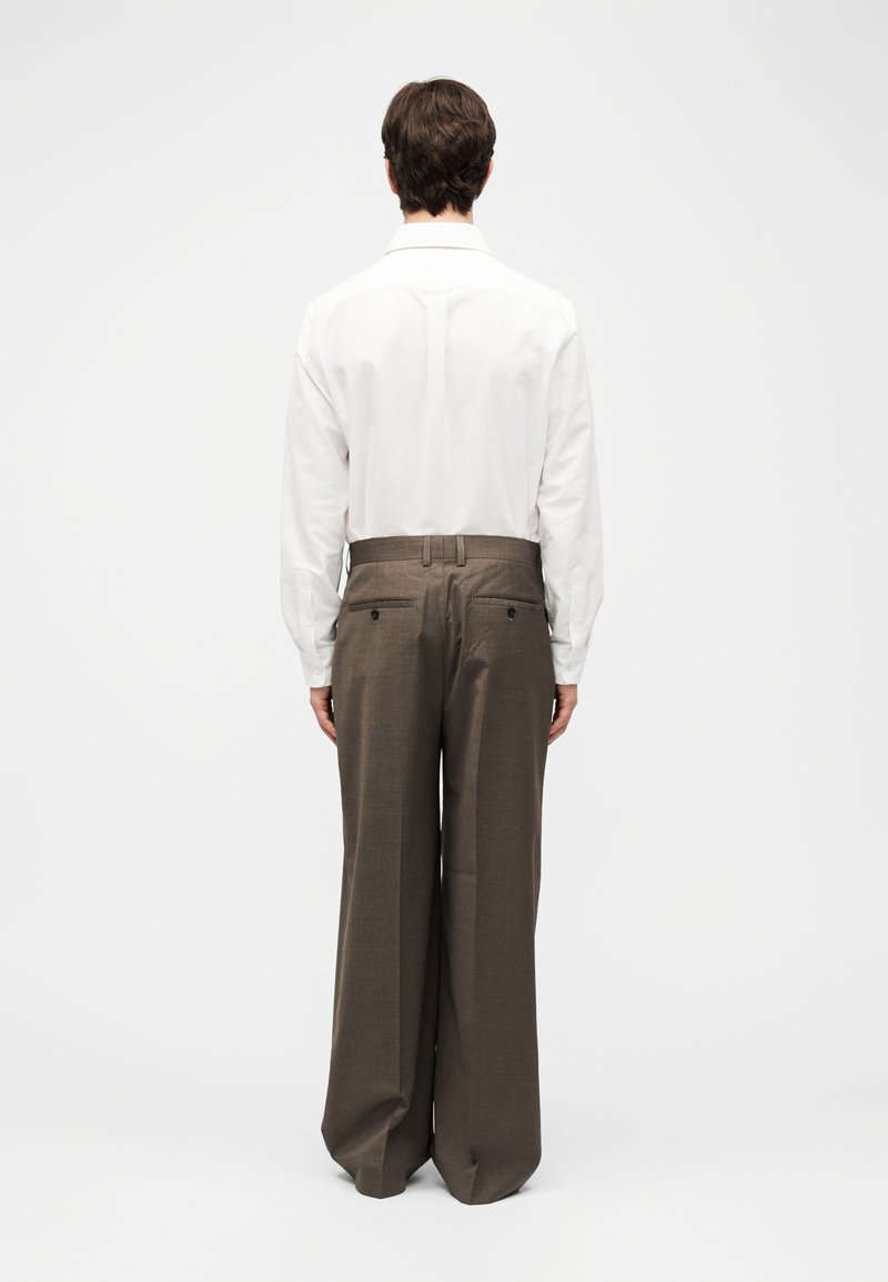 Mann steht mit dem Rücken zur Kamera, trägt ein weißes langärmliges Hemd, das in eine braune Hose mit weitem Bein und Gesäßtaschen gesteckt ist.