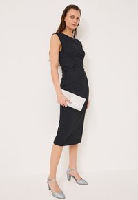 Svart ärmlös klänning med rynkade detaljer, knälång, matchad med en silverglittrande clutch och matchande låga silverklackar.