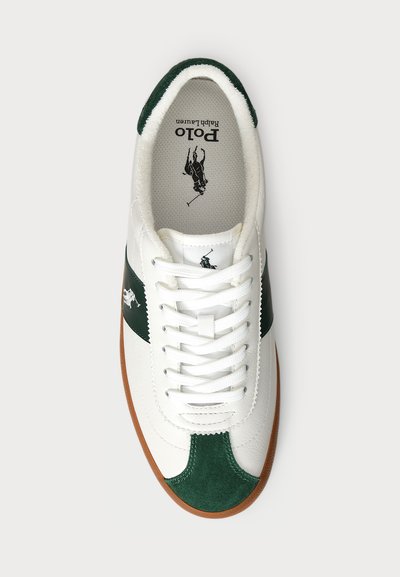 Vit sneaker med gröna mockadetaljer och ränder, strukturerat läderövre, platta vita snören och en brun gummisula. Innehåller logotyp inuti.
