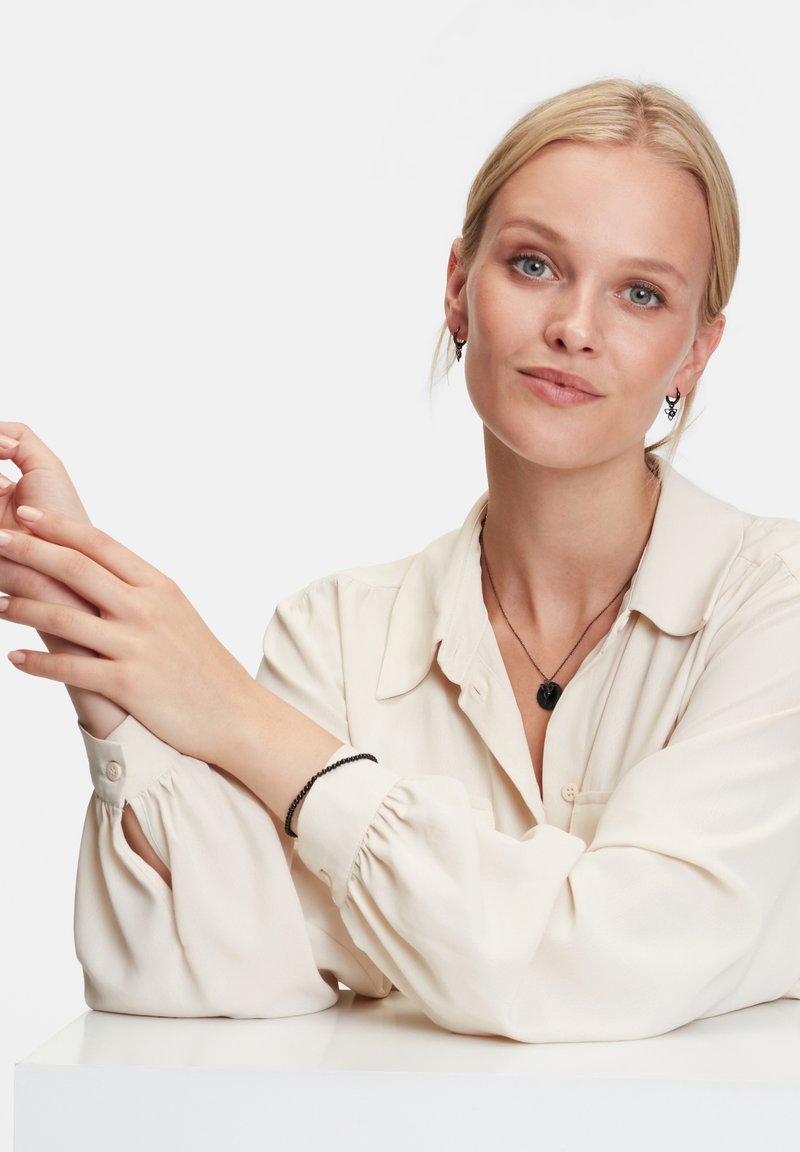 Zwarte juwelenset tegen een lichtblouson shirt. Het shirt heeft zachte, puffy mouwen. Een armband completeert de ketting en oorbellen.
