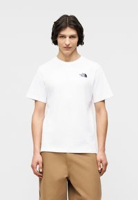 Νεαρός άνδρα ς με σκούρα κυματιστά μαλλιά που φοράει λευκό t-shirt The North Face και ανοιχτό καφέ παντελόνι, με φόντο ένα απλό ανοιχτό φωτεινό φόντο.