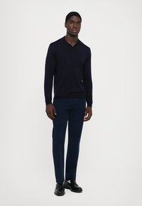 Young man standing wearing a navy long-sleeve polo shirt, dark blue pants, and black leather shoes against a plain white background.