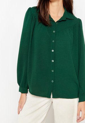 Femme portant un chemisier boutonné vert foncé à manches longues bouffantes et un pantalon blanc, debout devant un fond uni.