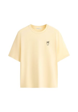 T-shirt jaune clair à manches courtes et col rond avec un petit contour noir représentant une boisson avec une paille et une garniture sur la poitrine gauche.