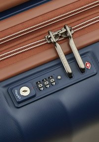 Valigia rigida blu navy con finitura testurizzata, doppi zip marroni, lucchetto combinato integrato e dettaglio con foro per chiave per un accesso sicuro.