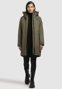 Olijfgroene parka met een met bont gevoerde capuchon, lange mouwen, een ritssluiting aan de voorkant, zijvakken en zwarte accenten. Draagt over een zwarte top en leggings.