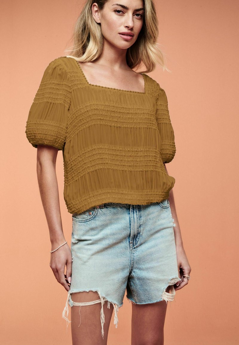 Top marrone con texture, scollatura quadrata, maniche a sbuffo e dettagli arricciati, abbinato a pantaloni corti in denim chiaro con orli sfilacciati.