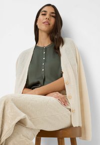Crème cardigan met knoopdetails over een donkergroene blouse. Beige textuur broek. Zittende pose, minimalistische achtergrond, houten kruk.