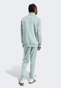 Svijetlozeleni trenirka izrađena od glatkog materijala, s bijelim bočnim prugama, visokim ovratnikom te rebrastim manžetama i pojasom.