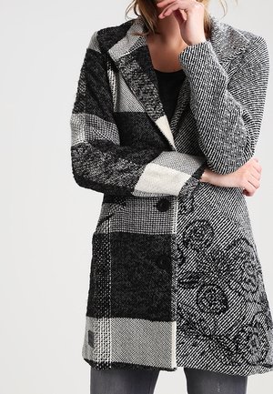 Vrouw met een zwart-witte wollen jas met patroon en knopen, staand met gekruiste armen en een hand bij het gezicht.