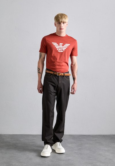 Red short-sleeve T-shirt with a white logo, paired with dark jeans and white sneakers. The model has a slender build and tattoos.
