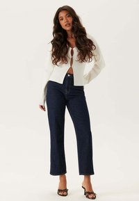 Vit långärmad cardigan med ett nyckelhålsdetalj fram, kombinerad med hög midja, mörkblå raka jeans och svarta klacksandaler.