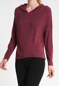 Burgundy long-sleeve hoodie with a v-neck and relaxed fit. Soft fabric with dropped shoulders. Paired with black pants.