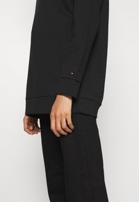 Černá mikina a kalhoty s volným střihem. Mají žebrovaný lem a manžety, na rukávu je malý barevný logo akcent.