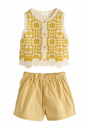 Háčkovaná bezrukávová vesta so žlto-krémovým kvetinovým vzorom a drevenými gombíkmi, v kombinácii so žltými krátkymi nohavicami s gumičkou v páse a kockovaným vzorom.