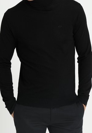 Homme portant un pull à col roulé noir ajusté et un pantalon noir, les mains partiellement dans les poches, sur un fond blanc uni.